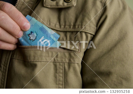 Hand putting Canadian money in pocket.person placing a blue Canadian five-dollar banknote into the chest pocket of a khaki jacket, symbolizing savings, cash handling, or financial security. 132935258