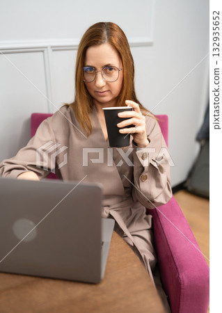 Businesswoman remote work coffee break using laptop in stylish modern office chair Businesswoman remote work coffee break using laptop in stylish modern office chair 132935652