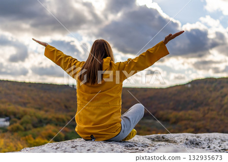 Woman nature freedom sitting on cliff rock with arms raised admiring the cloudy sky 132935673