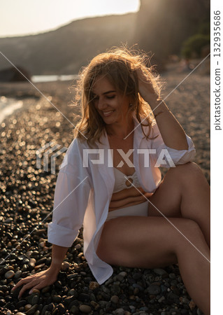 Woman beach sunset. Young woman sitting on pebble beach at sunset, smiling with golden light in her hair, enjoying peaceful evening by the sea Woman beach sunset. Young woman sitting on pebble beach at sunset, smiling with golden light in her hair, enjoying peaceful evening by the sea 132935686