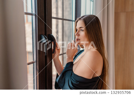 Woman, window, contemplation. Young female stands by window, touching lips in a thoughtful pose at home. 132935706