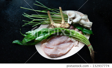 Top view of material for made Tom Kha Kai. With Raw chicken, galangal, lemongrass, kaffir lime leaves and mushroom. Arranged on a plate on a black background. Top view of material for made Tom Kha Kai. With Raw chicken, galangal, lemongrass, kaffir lime leaves and mushroom. Arranged on a plate on a black background. 132937774