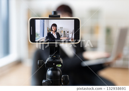 Caucasian woman in business attire recording herself on smartphone discussing business topics in office environment. Concept of video blogging, online content creation. Caucasian woman in business attire recording herself on smartphone discussing business topics in office environment. Concept of video blogging, online content creation. 132938691
