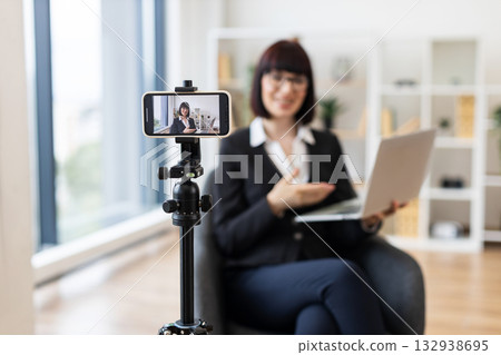 Caucasian businesswoman in her 30s filming vlog content with smartphone and laptop on desk, engaging with her audience in modern professional workplace. Caucasian businesswoman in her 30s filming vlog content with smartphone and laptop on desk, engaging with her audience in modern professional workplace. 132938695