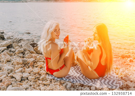 Watermelon Beach Summer: Two women enjoy refreshing watermelon slices on a rocky beach during a sunny day. 132938714