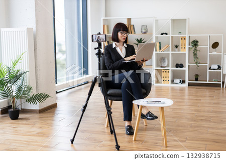 Caucasian businesswoman in modern office filming content on tripod camera holding laptop, delivering professional message. Bright interior with natural light highlights detailed setting Caucasian businesswoman in modern office filming content on tripod camera holding laptop, delivering professional message. Bright interior with natural light highlights detailed setting 132938715