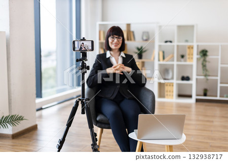 Caucasian adult woman using camera for recording business-related content in modern office environment. Focus on creating professional material remotely with digital tools. 132938717