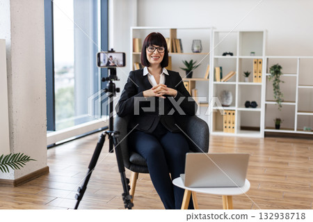 Caucasian adult woman using camera for recording business-related content in modern office environment. Focus on creating professional material remotely with digital tools. Caucasian adult woman using camera for recording business-related content in modern office environment. Focus on creating professional material remotely with digital tools. 132938718