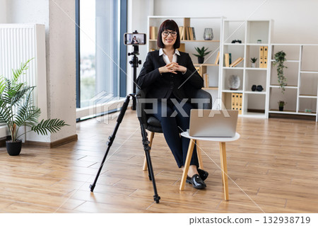 Caucasian woman aged 30-40 recording business vlog through camera in modern office. Professional attire shows corporate setting. Bright room features creative workspace environment. 132938719