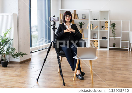 Caucasian woman aged 30-40 recording business vlog through camera in modern office. Professional attire shows corporate setting. Bright room features creative workspace environment. Caucasian woman aged 30-40 recording business vlog through camera in modern office. Professional attire shows corporate setting. Bright room features creative workspace environment. 132938720