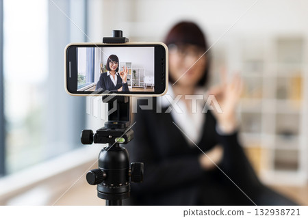 Caucasian woman wearing formal attire recording video blog using smartphone on tripod in modern office setting. Concept themes include business networking, educational content creation. Caucasian woman wearing formal attire recording video blog using smartphone on tripod in modern office setting. Concept themes include business networking, educational content creation. 132938721