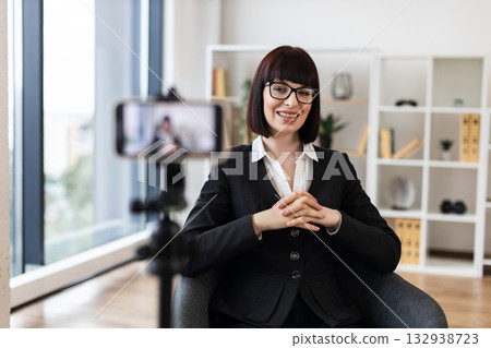 Caucasian woman in early 30s in business suit recording video content in office setting. Concept includes leadership, modern technology, and communication. Scene portrays professionalism. 132938723