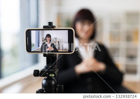 Caucasian woman in early 30s in business suit recording video content in office setting. Concept includes leadership, modern technology, and communication. Scene portrays professionalism. Caucasian woman in early 30s in business suit recording video content in office setting. Concept includes leadership, modern technology, and communication. Scene portrays professionalism. 132938724