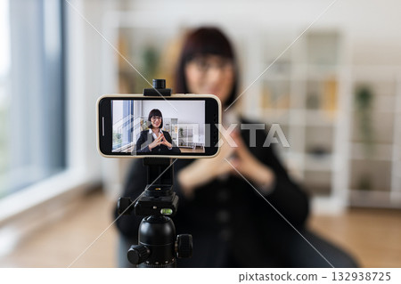 Caucasian woman recording video blog about business concepts and ideas using smartphone camera. Shows creative workspace with natural lighting for content creation and technology usage. 132938725