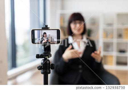 Caucasian woman recording video blog about business concepts and ideas using smartphone camera. Shows creative workspace with natural lighting for content creation and technology usage. Caucasian woman recording video blog about business concepts and ideas using smartphone camera. Shows creative workspace with natural lighting for content creation and technology usage. 132938730