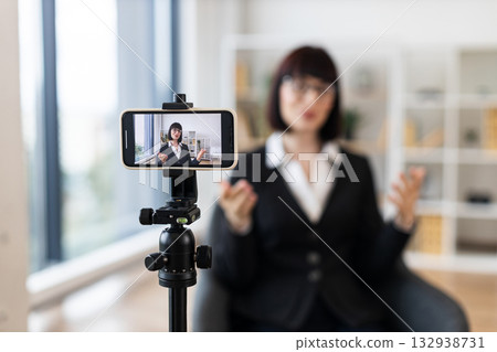 Caucasian woman recording video blog about business topics using smartphone camera on tripod. Professional indoor office setup emphasizes modern technology, content creation, and professionalism. Caucasian woman recording video blog about business topics using smartphone camera on tripod. Professional indoor office setup emphasizes modern technology, content creation, and professionalism. 132938731
