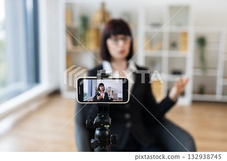 Caucasian woman recording video blog indoors with smartphone on tripod, sharing business insights and advice. She has dark hair, glasses, wearing formal clothing. 132938745