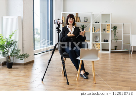 Caucasian woman recording video blog on business topics sitting in stylish office interior. Professional setup includes camera, tripod, laptop demonstrating work focus and modern office aesthetic. Caucasian woman recording video blog on business topics sitting in stylish office interior. Professional setup includes camera, tripod, laptop demonstrating work focus and modern office aesthetic. 132938758