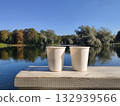 Two white paper cups are placed on a wooden table, with a view of a lake and a park. Picturesque nature. A cozy outdoor recreation. Gatchina Palace Park. Russia. 132939566
