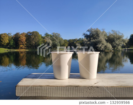 Two white paper cups are placed on a wooden table, with a view of a lake and a park. Picturesque nature. A cozy outdoor recreation. Gatchina Palace Park. Russia. Two white paper cups are placed on a wooden table, with a view of a lake and a park. Picturesque nature. A cozy outdoor recreation. Gatchina Palace Park. Russia. 132939566