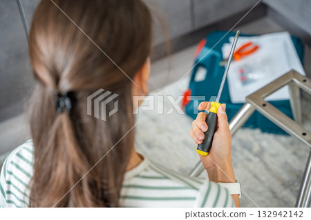 Young woman seen from behind assembling a chair with screwdrivers on the floor at home. Feminist concept of female independence and do it yourself furniture assembly. 132942142
