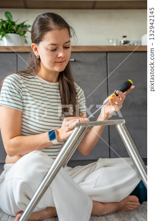 Smiling young woman assembling a chair with tools on the floor. Concept of joy and satisfaction from independent home projects. 132942145