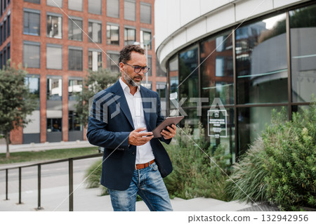 Businessman in front of office building, holding tablet and reading something. 132942956
