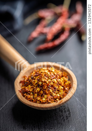 Red chili peppers flakes. Milled spicy peppers on wooden scoop on black table. Red chili peppers flakes. Milled spicy peppers on wooden scoop on black table. 132943072