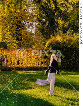 Happy young blonde woman wearing casual clothes is walking and slightly dancing in park during beautiful sunny autumn day, with colorful foliage in background. Photoshoot of happy woman in autumn park Happy young blonde woman wearing casual clothes is walking and slightly dancing in park during beautiful sunny autumn day, with colorful foliage in background. Photoshoot of happy woman in autumn park 132943310