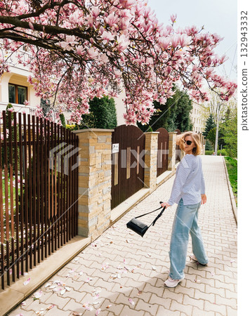 Stylish woman walking along sidewalk, surrounded by vibrant blooms of magnolia tree, savoring beauty of sunny spring day while carrying chic black purse. Blonde woman walking under magnolia blossoms 132943332