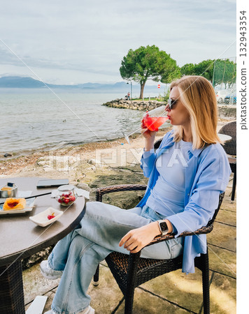 Young blonde woman sitting at table by Lake Garda, drinking red cocktail and enjoying view of lake and mountains during relaxing vacation. Blonde woman relaxing at Lake Garda table, sipping cocktail Young blonde woman sitting at table by Lake Garda, drinking red cocktail and enjoying view of lake and mountains during relaxing vacation. Blonde woman relaxing at Lake Garda table, sipping cocktail 132943354