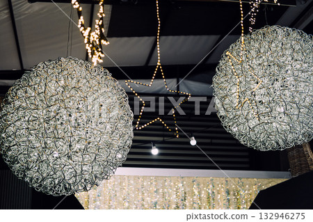 Illuminated wire-frame stars and wreaths hanging from a ceiling in a modern holiday display. Christmas lighting, geometric festive decor, sculptural illumination, contemporary holiday styling Illuminated wire-frame stars and wreaths hanging from a ceiling in a modern holiday display. Christmas lighting, geometric festive decor, sculptural illumination, contemporary holiday styling 132946275