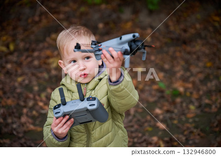 A curious young boy holds a drone in one hand and a remote controller in the other, exploring nature among colorful autumn leaves. His excitement fills the air. A curious young boy holds a drone in one hand and a remote controller in the other, exploring nature among colorful autumn leaves. His excitement fills the air. 132946800