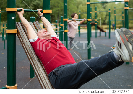 Senior man goes in for sports in city park on sports ground - doing exercises to pull his legs to chest Senior man goes in for sports in city park on sports ground - doing exercises to pull his legs to chest 132947225