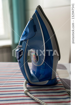 Blue electric iron positioned on striped fabric surface, showcasing modern design and functionality, ideal for home appliance and household tasks, with vibrant colors enhancing visual appeal Blue electric iron positioned on striped fabric surface, showcasing modern design and functionality, ideal for home appliance and household tasks, with vibrant colors enhancing visual appeal 132947541