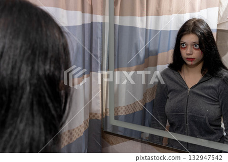 Young woman with dark hair, wearing a gray hoodie, gazes into a mirror with dramatic makeup, creating a haunting atmosphere in a bathroom setting with patterned curtains Young woman with dark hair, wearing a gray hoodie, gazes into a mirror with dramatic makeup, creating a haunting atmosphere in a bathroom setting with patterned curtains 132947542