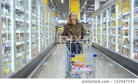 Young lady maneuvering a shopping cart down the frozen food aisle in a contemporary grocery store, browsing for groceries 132947543