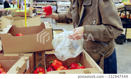 Woman in a leather jacket picks bell peppers from cardboard boxes in a grocery store, while opening a plastic bag 132947549