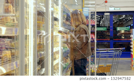 Grocery shopping routine, Woman choosing frozen meals carefully, Urban shopper selecting groceries with intent, Customer in city supermarket methodically picks frozen food items for weekly 132947582