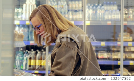 Young woman with glasses carefully selecting refrigerated products in a supermarket, reading labels and comparing choices Young woman with glasses carefully selecting refrigerated products in a supermarket, reading labels and comparing choices 132947592