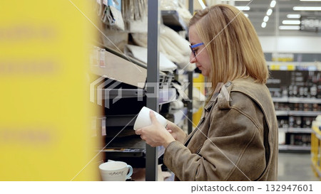 Young woman carefully selects a white coffee mug in a home goods store before making her decision Young woman carefully selects a white coffee mug in a home goods store before making her decision 132947601