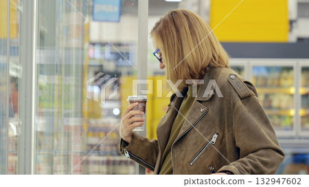 Woman enjoys shopping alone in a supermarket, sipping coffee while selecting groceries from the refrigerated aisle. She carefully reads labels and decides on fresh products 132947602