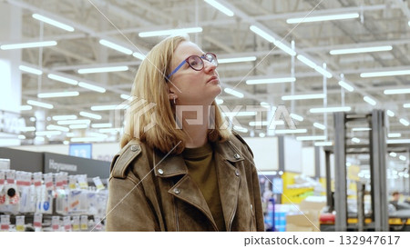 Young woman in glasses and a brown leather jacket gazes up at the ceiling lights in a spacious hardware store, possibly considering a purchase or admiring the store's design 132947617