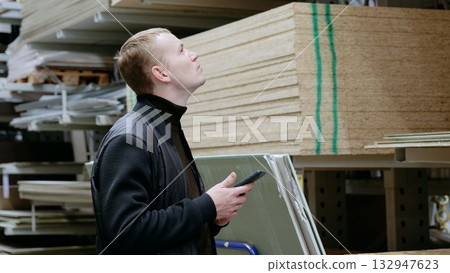 Customer is using a smartphone to compare construction materials for home renovation in a diy store. A man in a hardware store, choosing lumber, shelves with lumber. 132947623