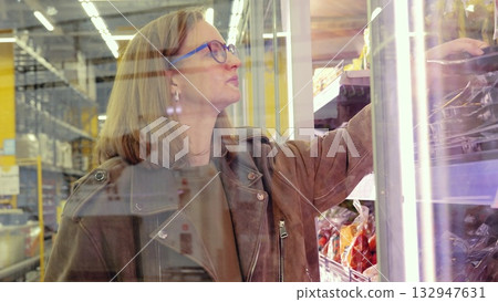Supermarket label inspection, Woman analyzing nutrition labels through refrigerated glass, Woman in coat and glasses carefully checks frozen food nutritional information through freezer door 132947631