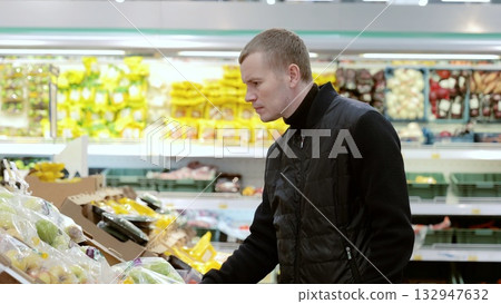 Man choosing fruits in supermarket, shelf with apples. Young man in casual winter attire carefully chooses healthy fruits and vegetables at the supermarket Man choosing fruits in supermarket, shelf with apples. Young man in casual winter attire carefully chooses healthy fruits and vegetables at the supermarket 132947632