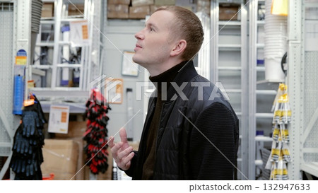 Customer makes a purchase decision while scanning the high shelves in a hardware store, considering a variety of products. A man in a hardware store choosing a tool. Customer makes a purchase decision while scanning the high shelves in a hardware store, considering a variety of products. A man in a hardware store choosing a tool. 132947633