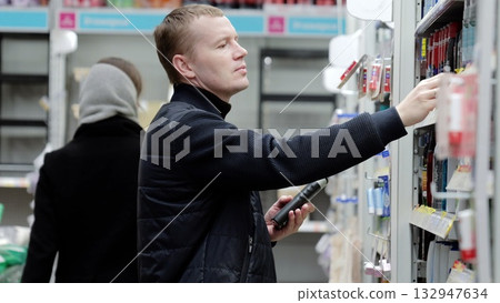 Man in a supermarket using a smartphone app to compare products while shopping, with blurred background of her walking. A man chooses hygiene products in a supermarket, shelves with shampoos Man in a supermarket using a smartphone app to compare products while shopping, with blurred background of her walking. A man chooses hygiene products in a supermarket, shelves with shampoos 132947634