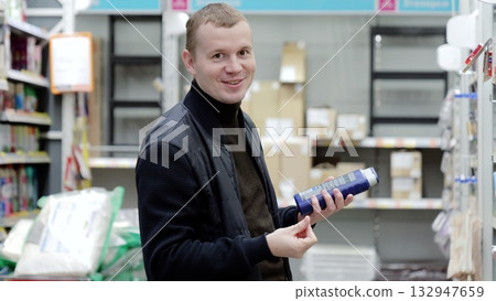 Man happily compares hair care products in a supermarket aisle, reading labels and ingredients with dedication. Background is blurred, creating cozy shopping atmosphere. Man happily compares hair care products in a supermarket aisle, reading labels and ingredients with dedication. Background is blurred, creating cozy shopping atmosphere. 132947659