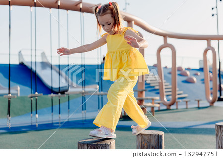 Little girl balancing on playground stumps, focused and active on a sunny day. 132947751